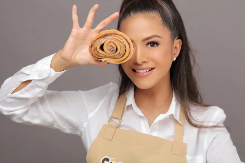 Woman in baker's apron holding a spiral pastry near her face, smiling